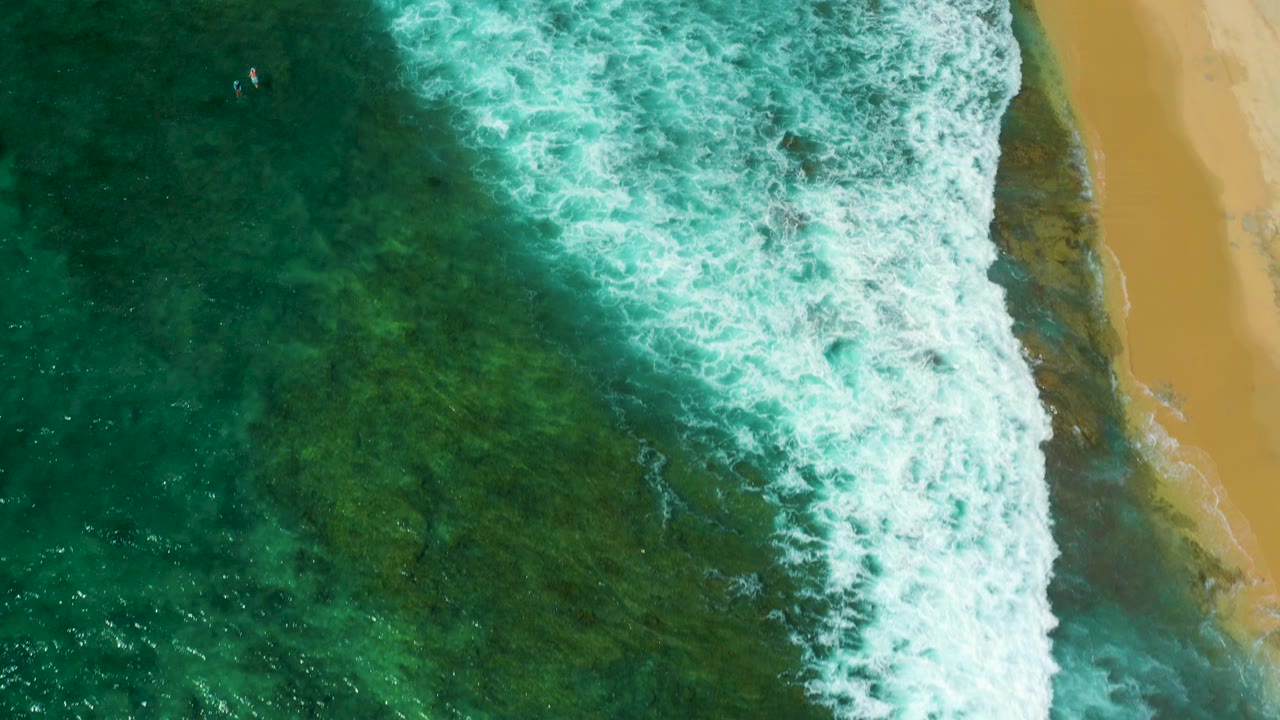 Overhead Shipwrecks Beach view with surf pushed farther across the frame, clear green water below, sand at the right margin, and no visible people.
