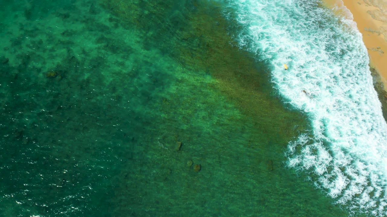 Overhead view of Shipwrecks Beach showing a broken white wave band along the right side, clear green water across the frame, small dark underwater shapes, and no visible people.