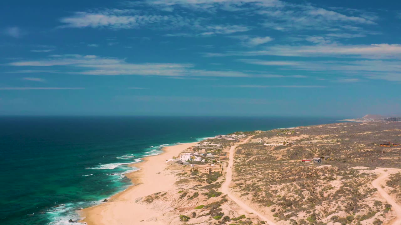 Aerial frame from Shipwrecks Beach showing breaking waves along the left edge, a broad sandy beach, low dunes, scattered scrub, and no visible people.