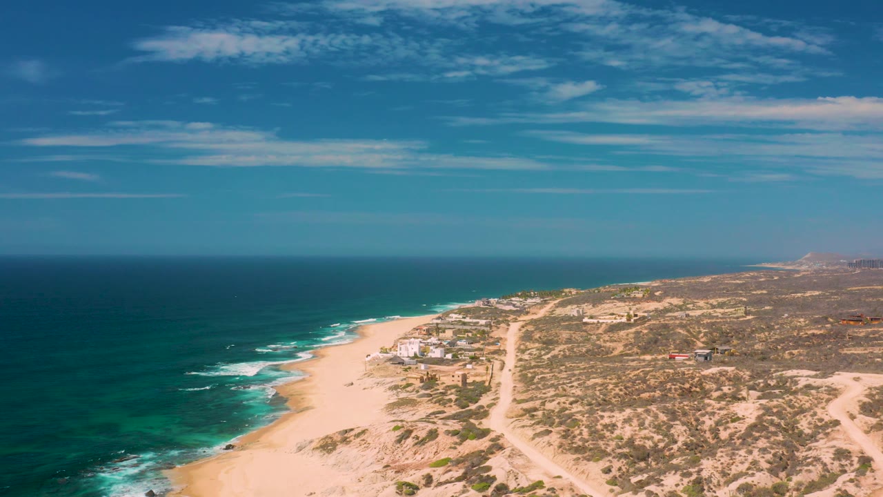 Aerial view over Shipwrecks Beach with the surf line on the left, a sandy road cutting through scrubland on the right, small distant structures, and no visible people.