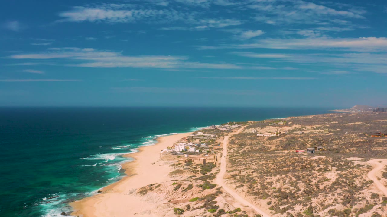 Elevated view of Shipwrecks Beach with white surf curling onto pale sand, open dunes in the foreground, scrubland inland, and no visible people.