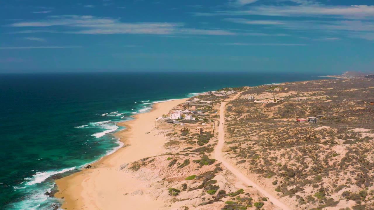 Aerial view at Shipwrecks Beach showing the sandy shoreline curving past dark blue water, small white wave breaks, scrub-covered dunes, distant low buildings, and no visible people.