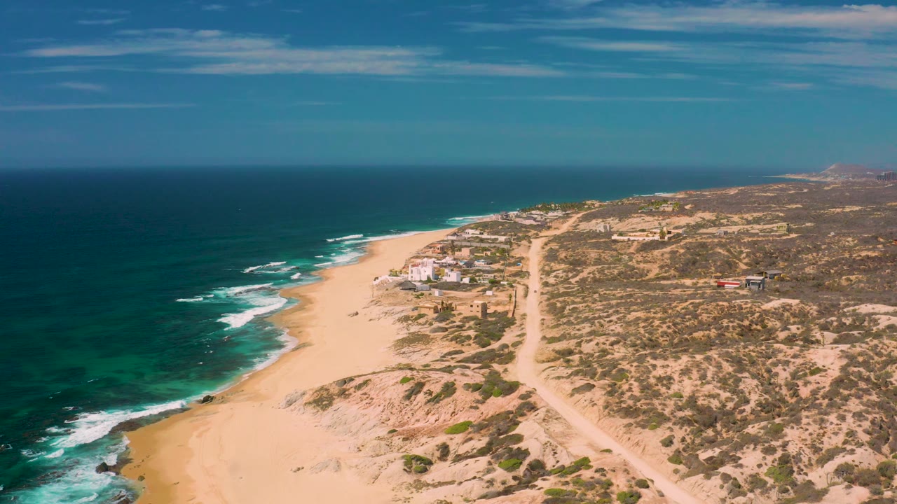 Shipwrecks Beach aerial frame with white surf bands along the left edge, pale sand through the middle, a straight dirt road inland, scattered low structures, hazy distant coastline, and no visible people.