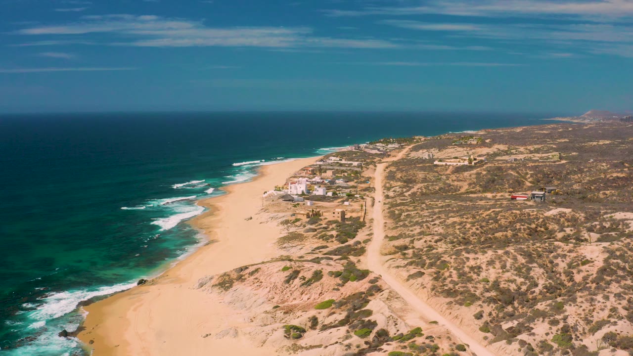 Wide drone-style view over Shipwrecks Beach with open sand in the lower center, dark green-blue water to the left, dry brush terrain to the right, a dirt road, low buildings, and no visible people.