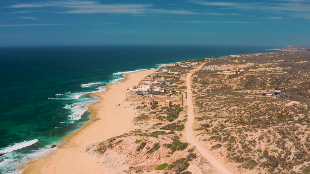 Wide aerial frame at Shipwrecks Beach in Baja California Sur, with darker blue water offshore, white breaks along the sand, a cluster of small coastal buildings, desert scrub, and no visible people.
