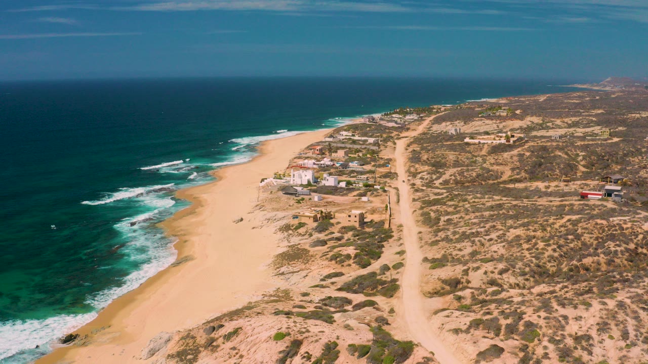 Opening aerial frame over Shipwrecks Beach in Baja California Sur, with dark blue ocean on the left, white surf, broad sand, scrub dunes, a sandy road, and no visible people.