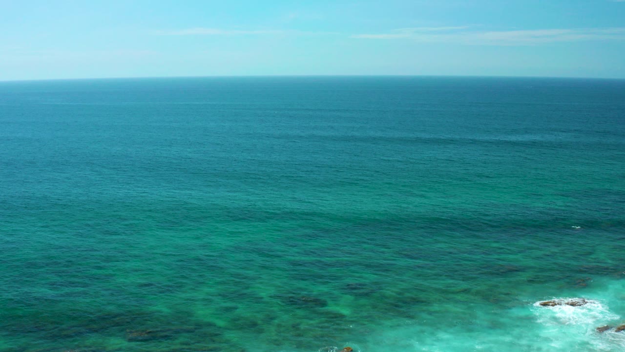 Wide aerial frame over Shipwrecks Beach, Baja California Sur, with turquoise water, darker reef bands, small rock breaks, a level horizon, and no visible people.