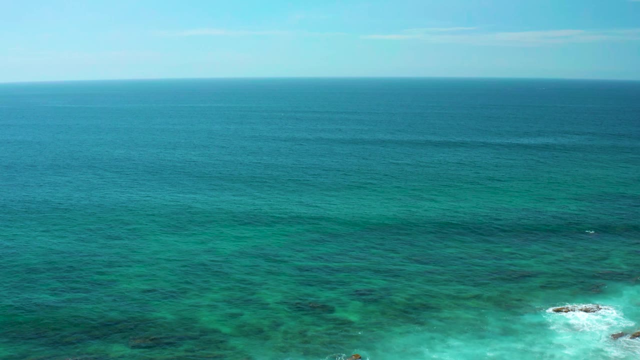 Aerial ocean view with turquoise shallows, darker water bands, small rocks, and white foam.