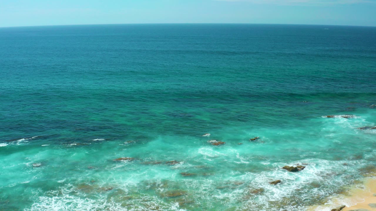 Aerial view of turquoise water and white surf over shallow rocks at Shipwrecks Beach, Baja California Sur, with deep blue ocean beyond and no visible people.