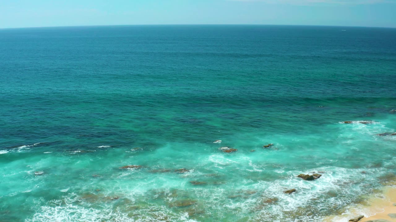 A high coastal view at Shipwrecks Beach shows blue-green water, scattered dark reef patches, white foam lines, and a narrow sand edge with no visible people.