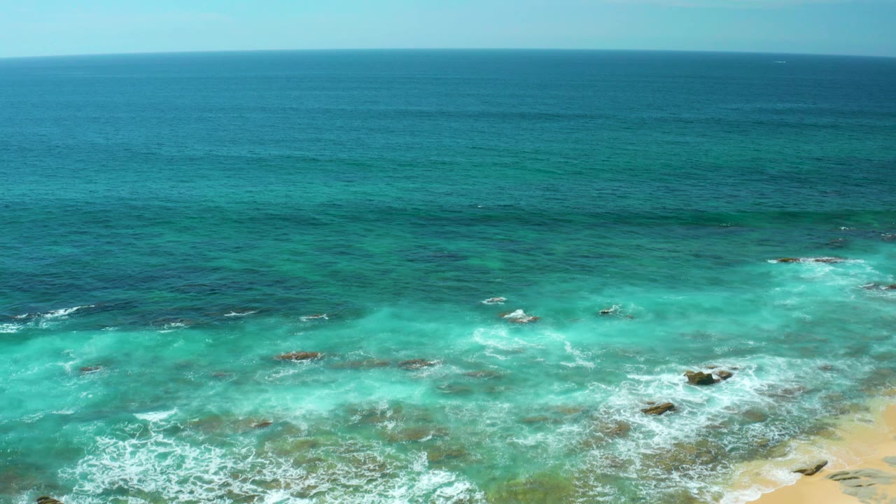 Turquoise surf spreads across rocky shallows at Shipwrecks Beach while a small strip of pale sand enters the lower right of the aerial frame, with no visible people.