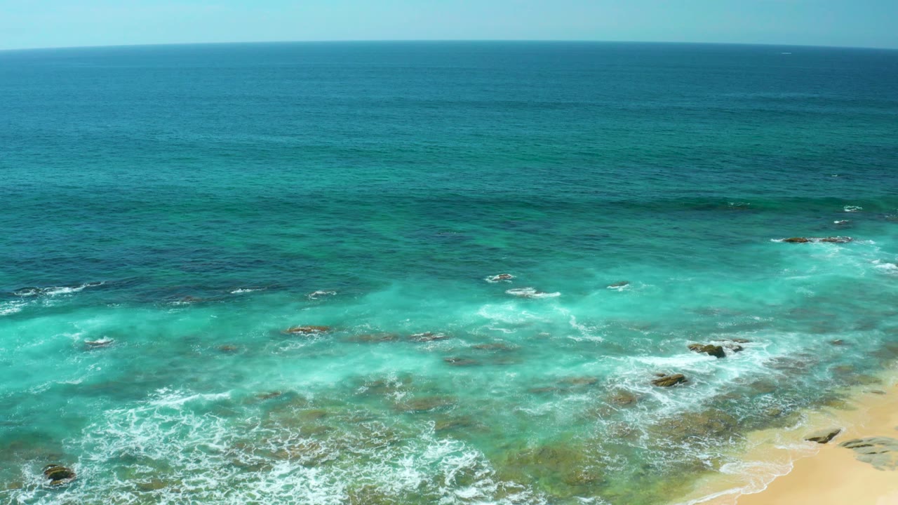 Aerial footage over Shipwrecks Beach shows clear cyan water, white breakers over rocks, a darker offshore band, and a sandy corner at the bottom right with no visible people.