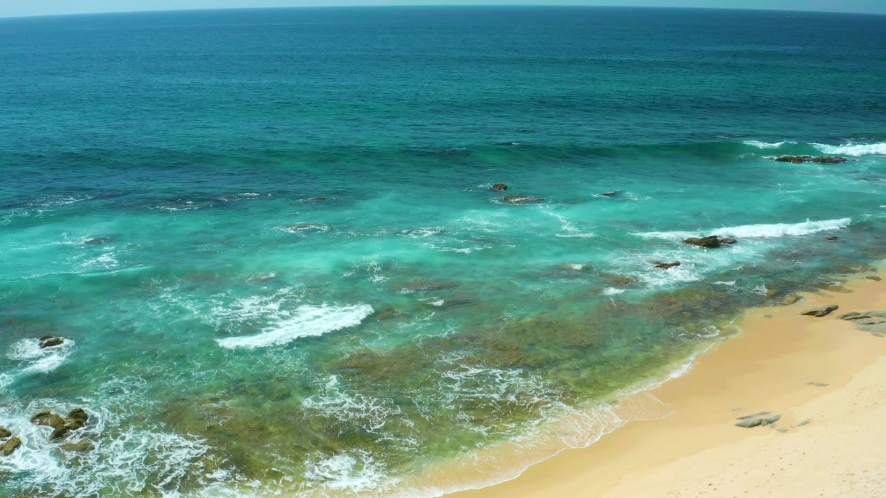 Foamy surf advances across shallow turquoise water at Shipwrecks Beach, with pale sand on the lower right and no visible people.