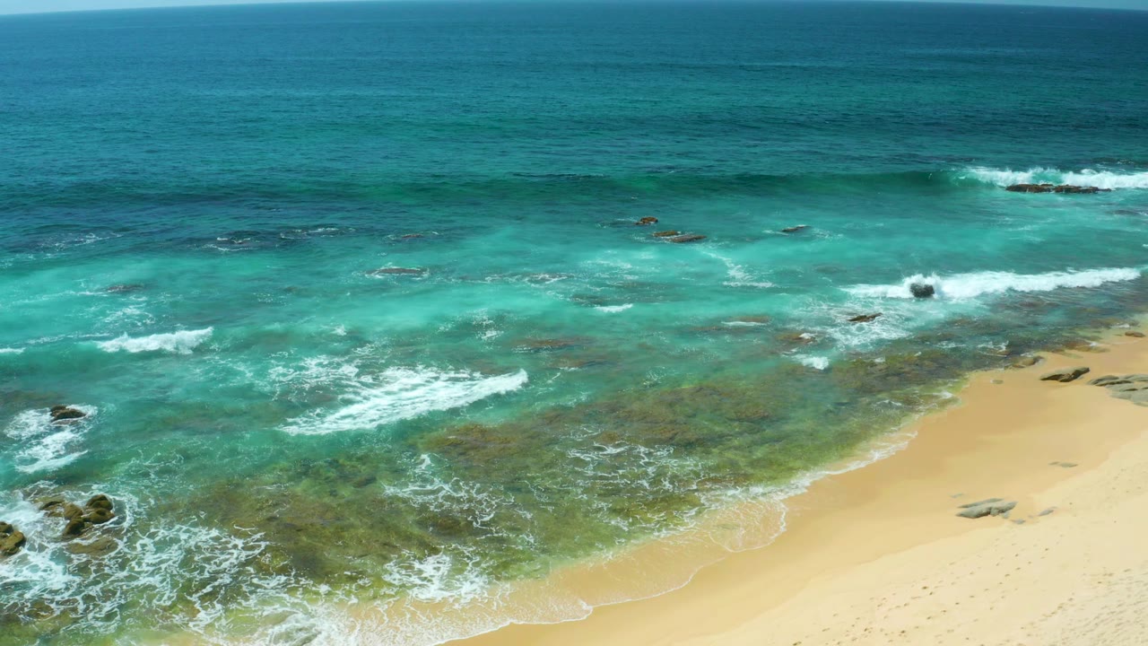 Clear surf breaks around scattered dark rocks near the sandy edge of Shipwrecks Beach, with open blue water beyond and no visible people.