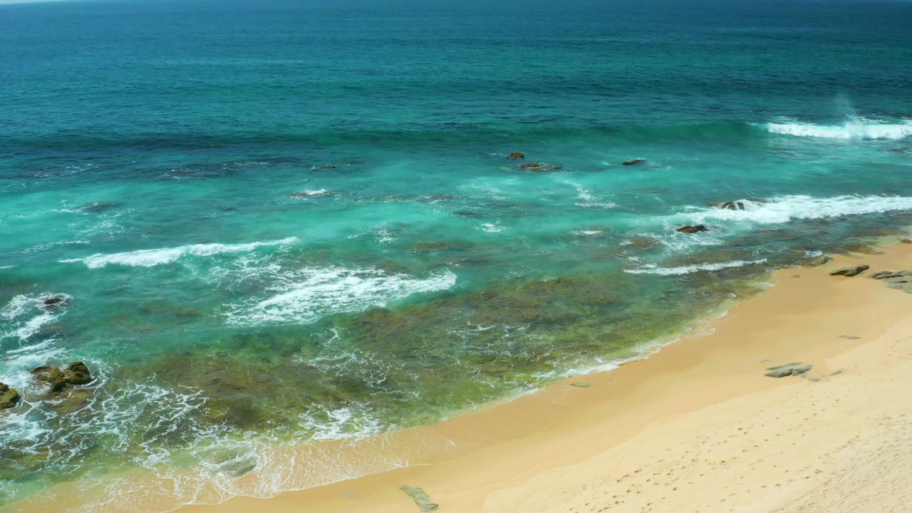 An elevated view of Shipwrecks Beach shows a broader band of sand below, surf streaks across the middle, dark rocks at left, and no visible people.