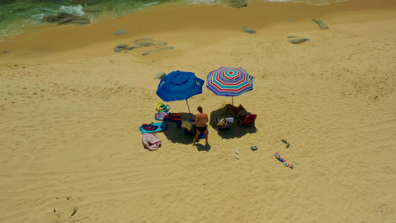 An overhead view at Shipwrecks Beach shows one adult standing beside the grouped umbrellas and chairs while the second person is partly obscured by shade and beach gear.