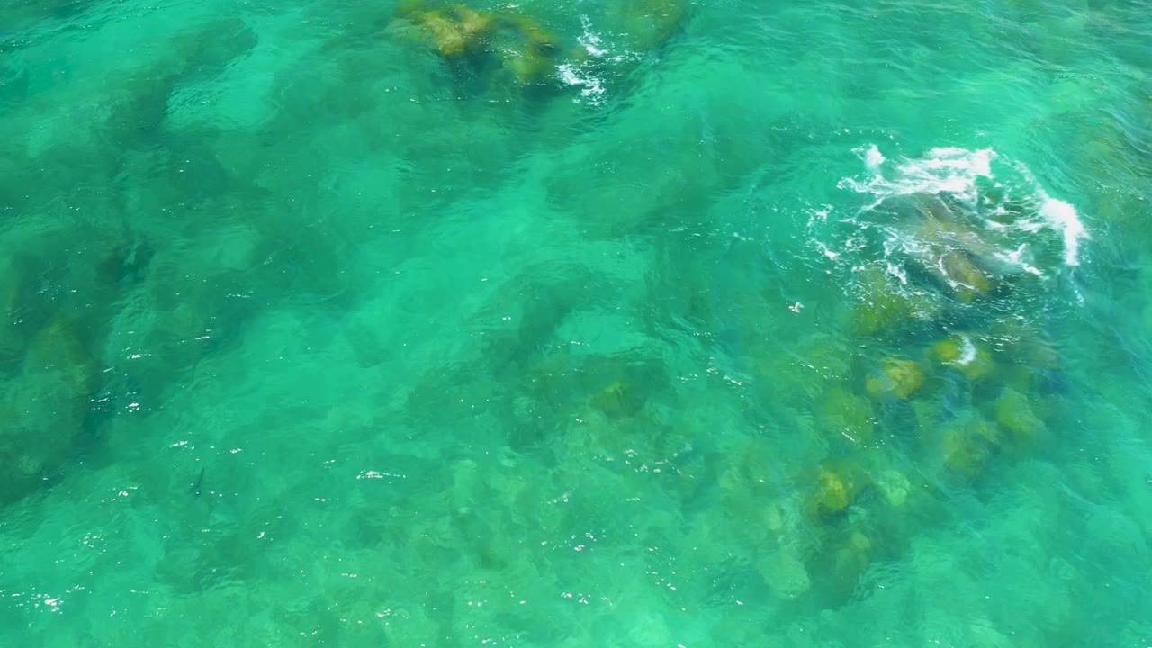 Overhead frame at Shipwrecks Beach showing a larger submerged rock cluster under blue-green water with a small white ripple breaking over it, no visible people.