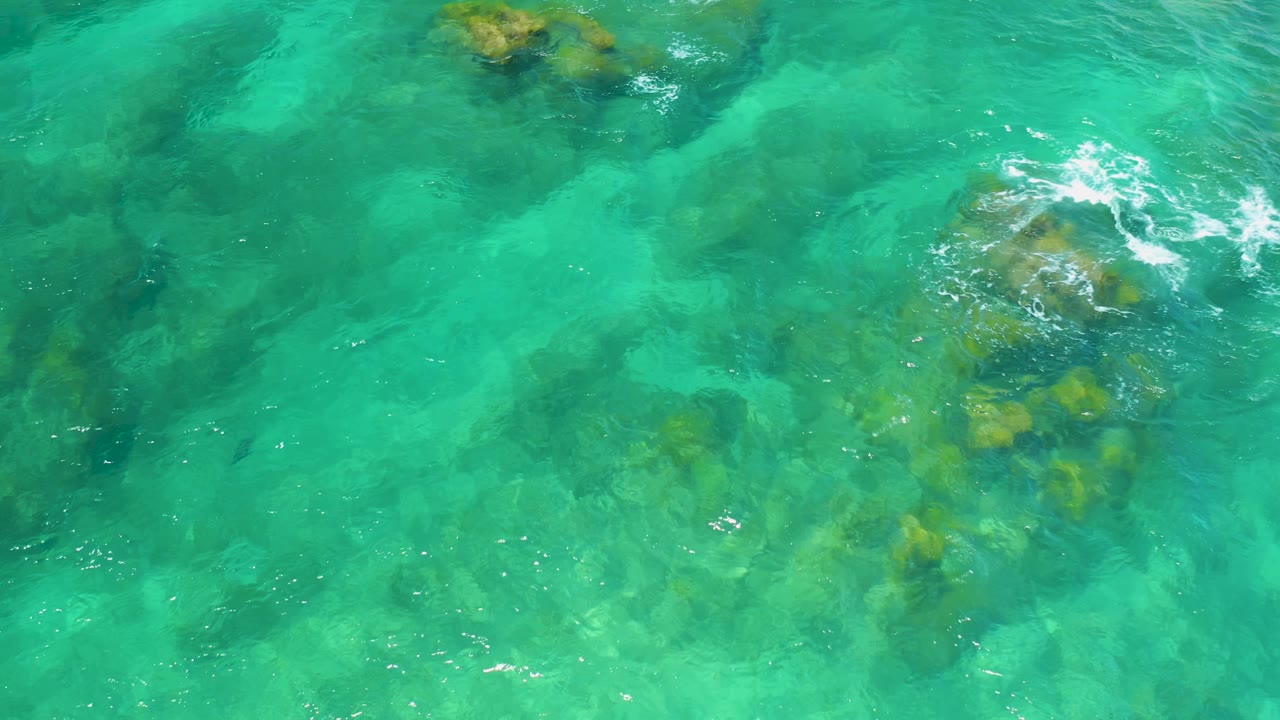 Overhead view of bright turquoise water at Shipwrecks Beach with a rounded rock group at upper right and scattered darker patches below the surface, no visible people.