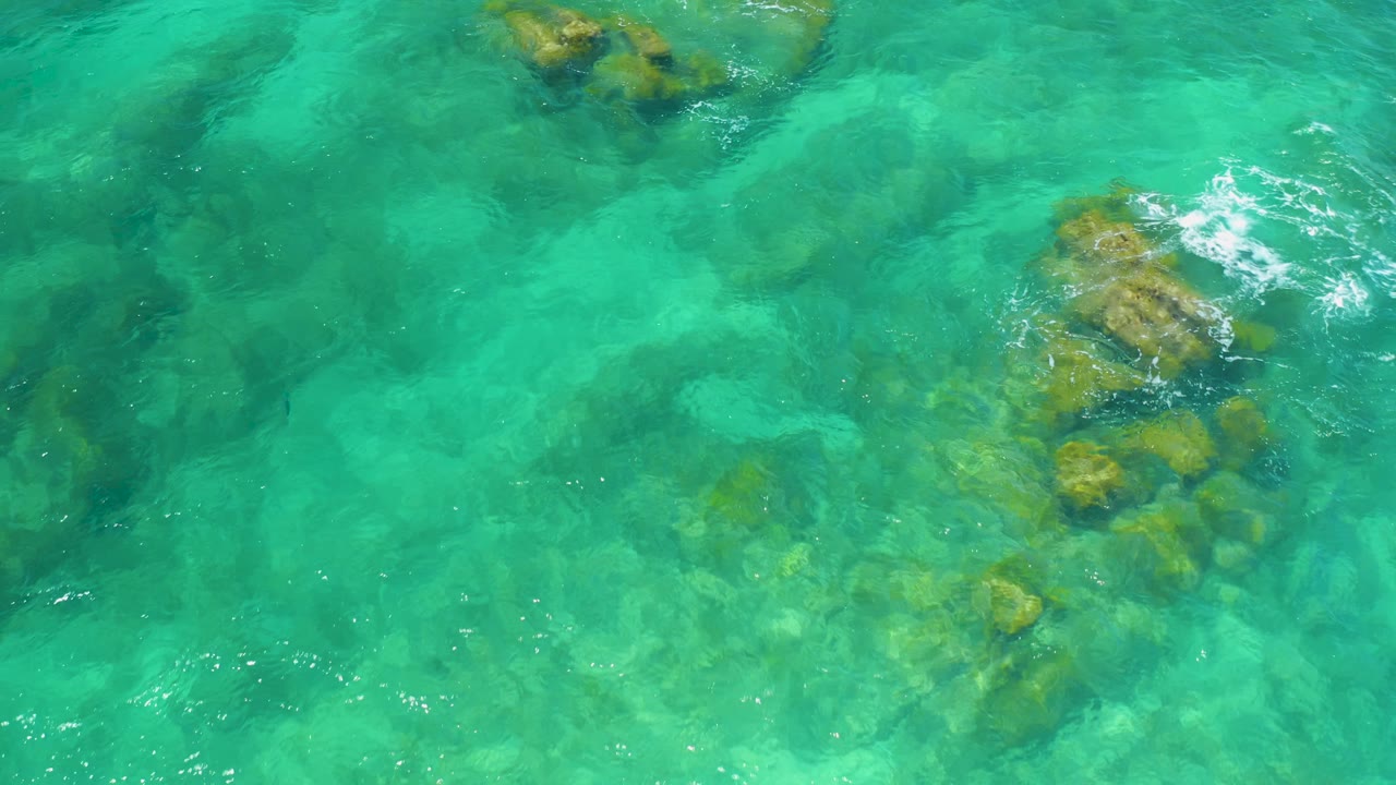 Overhead frame at Shipwrecks Beach with a submerged rock shelf entering from the top and a larger rock formation under calm teal water near center, no visible people.