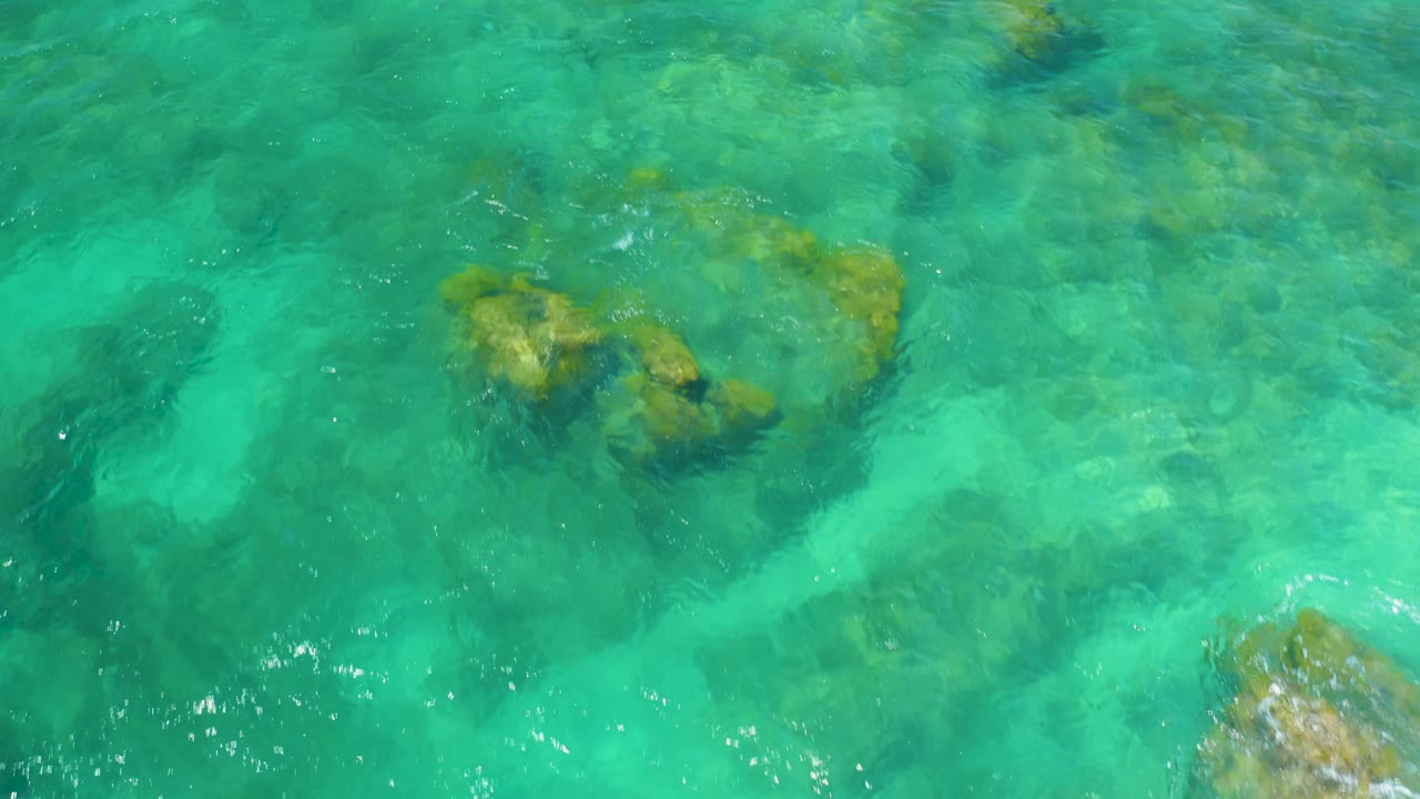 Aerial-looking top-down frame of Shipwrecks Beach water with a brown rock mass near the upper right and a brighter rock at the lower right edge, no visible people.