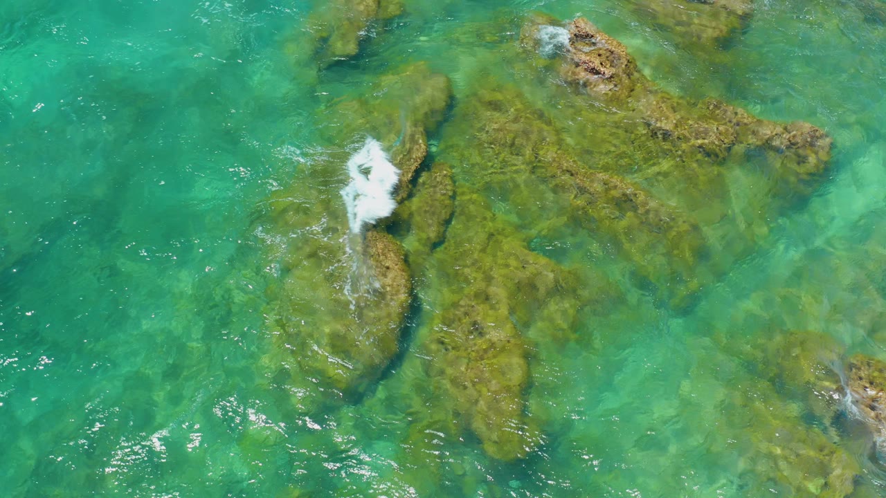 Overhead view at Shipwrecks Beach, Baja California Sur, showing green-blue water over moss-covered rocks with a small wave breaking near the left side and no visible people.