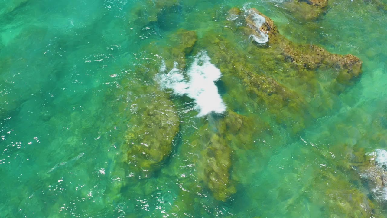 Aerial view at Shipwrecks Beach, Baja California Sur, where white surf spreads over shallow mossy rocks in clear green-blue water with no visible people.