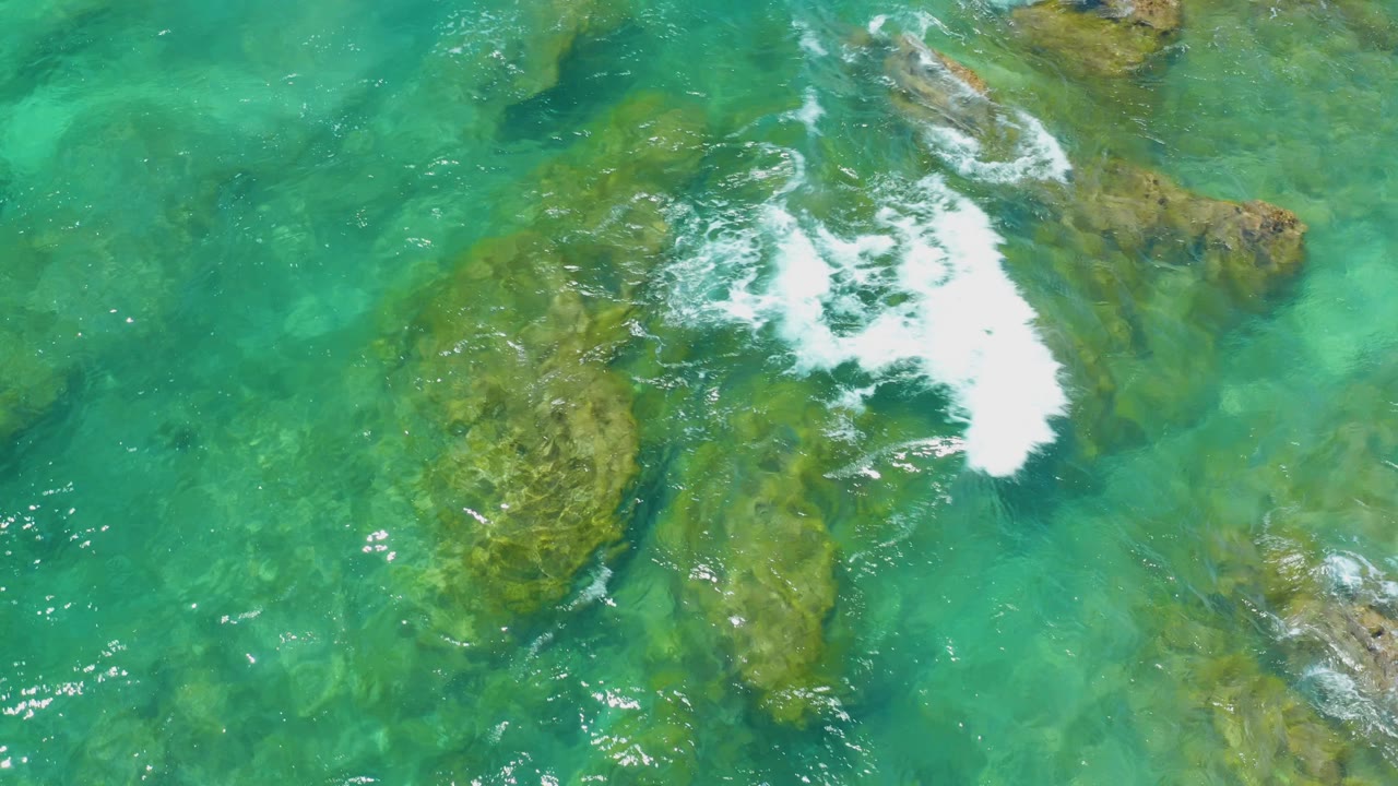 Overhead Shipwrecks Beach frame with a foamy wave breaking across the middle of shallow rock shelves, surrounded by turquoise water and no visible people.
