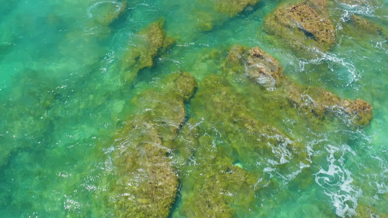 A top-down frame from Shipwrecks Beach shows a clustered rocky patch beneath turquoise water, with small white surf traces and no visible people.