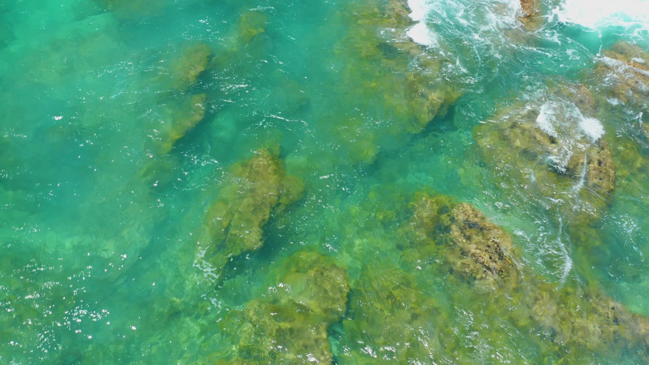 At Shipwrecks Beach in Baja California Sur, turquoise water covers scattered submerged rocks while white foam breaks along the upper edge, with no visible people.