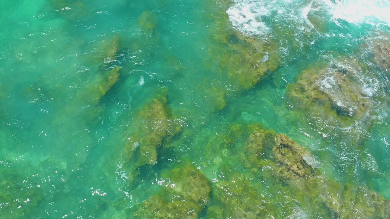 At Shipwrecks Beach in Baja California Sur, foamy surf spreads across the upper right while tan rocks remain visible through the turquoise shallows, with no visible people.