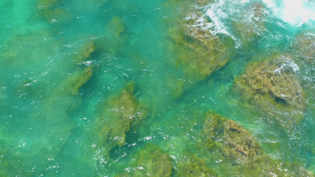 At Shipwrecks Beach in Baja California Sur, an overhead view shows clear turquoise water, scattered submerged rocks, and small white ripples across the surface, with no visible people.