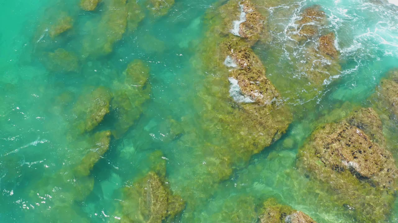 Aerial frame of green-blue water around uneven rock shelves at Shipwrecks Beach, Baja California Sur, with light foam along the upper rocks and no visible people.