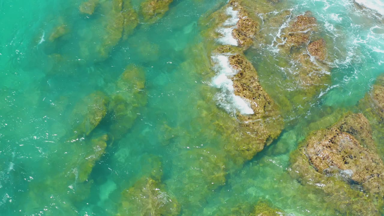 Top-down view at Shipwrecks Beach, Baja California Sur, where turquoise water covers shallow rocks and a brighter white break forms near the upper-right reef, with no visible people.