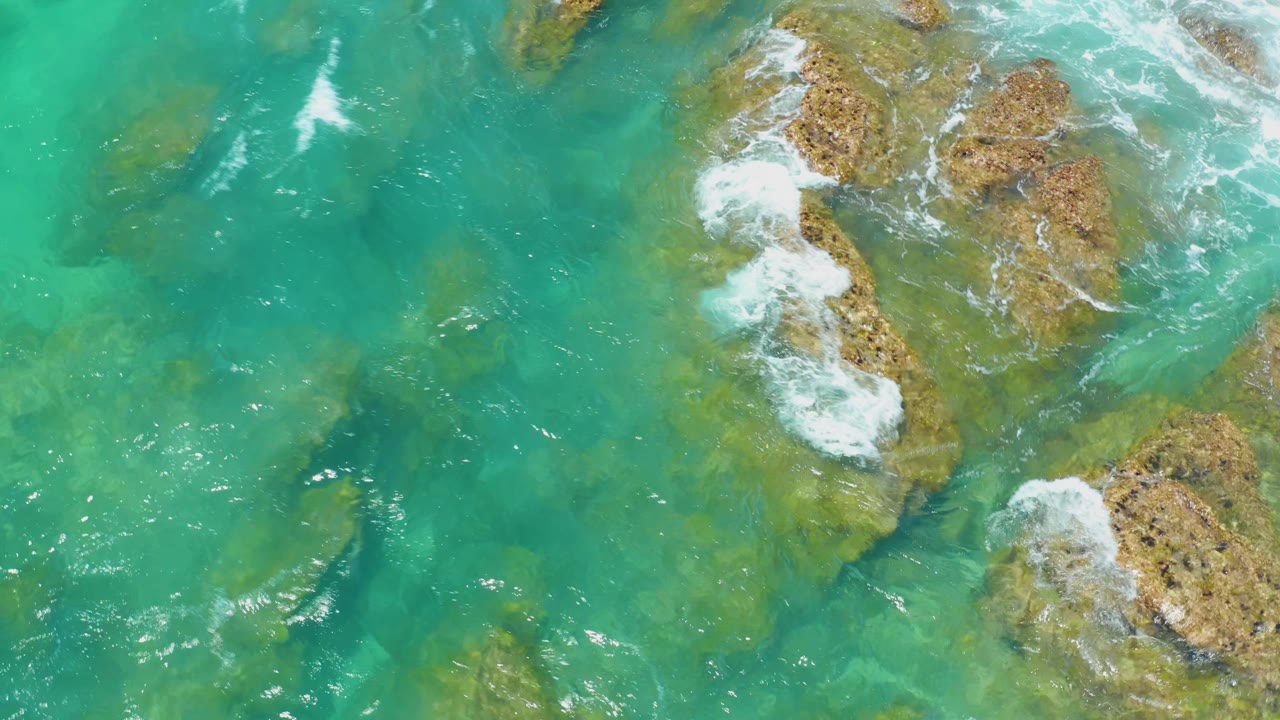 Overhead Shipwrecks Beach frame with white foam spreading across central brown rocks, clear turquoise water on both sides, and no visible people.