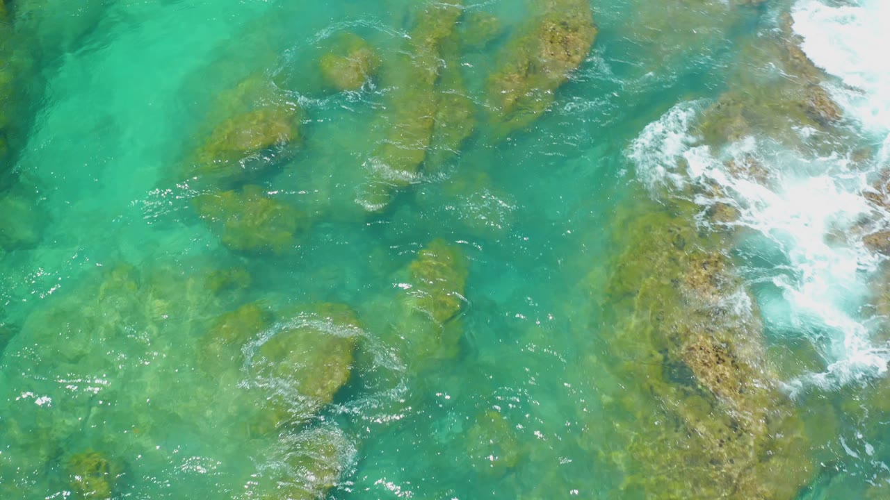 Top-down Shipwrecks Beach view with scattered shallow rocks under turquoise water, rippled highlights across the surface, white surf on the right, and no visible people.