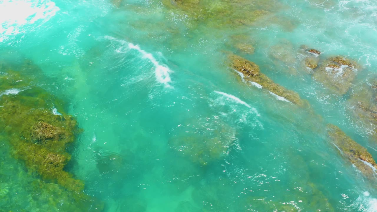 Overhead Shipwrecks Beach view with a white wave line entering from the upper left and reef patches visible through shallow turquoise water, no visible people.