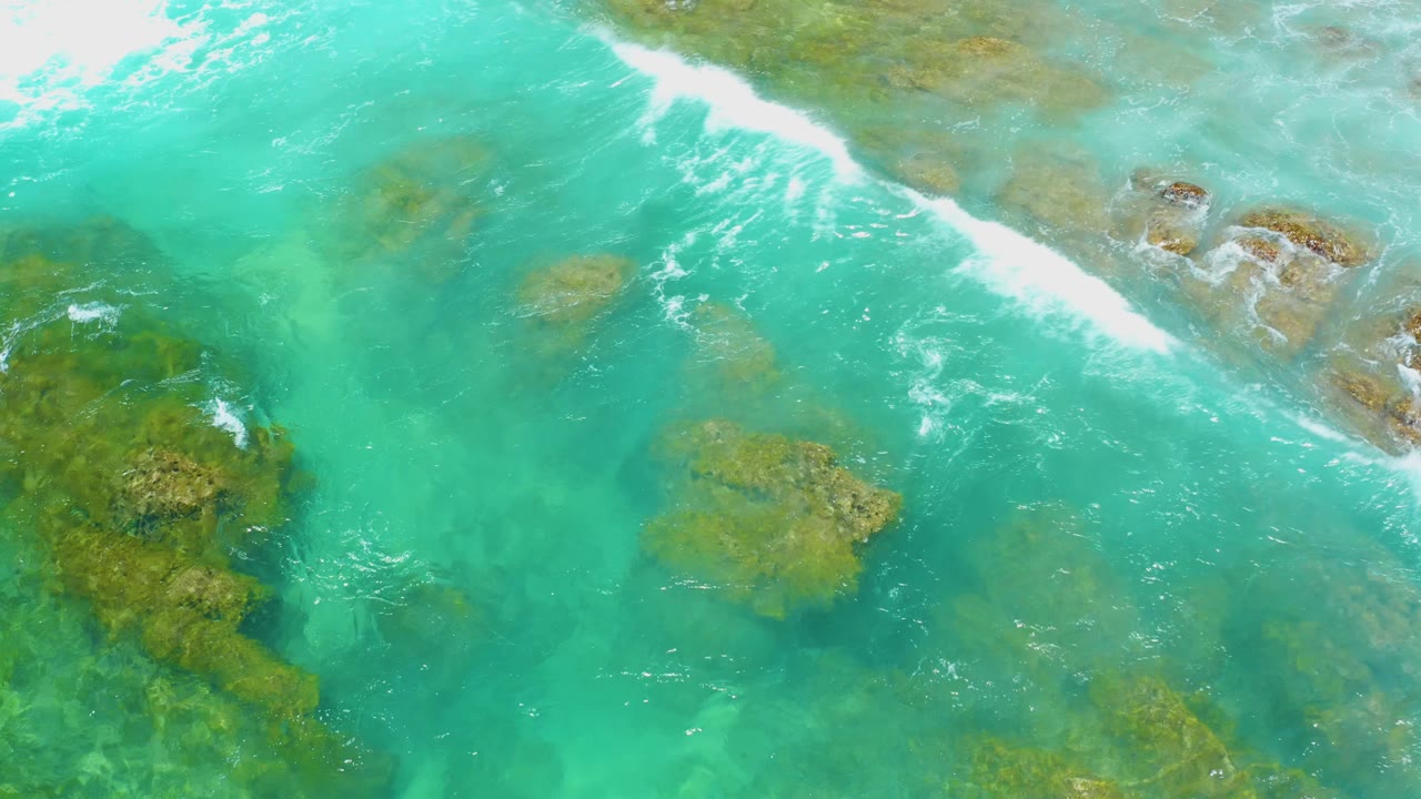 A diagonal band of white foam runs across shallow water at Shipwrecks Beach, with brown-green reef and rock shapes visible below, no visible people.