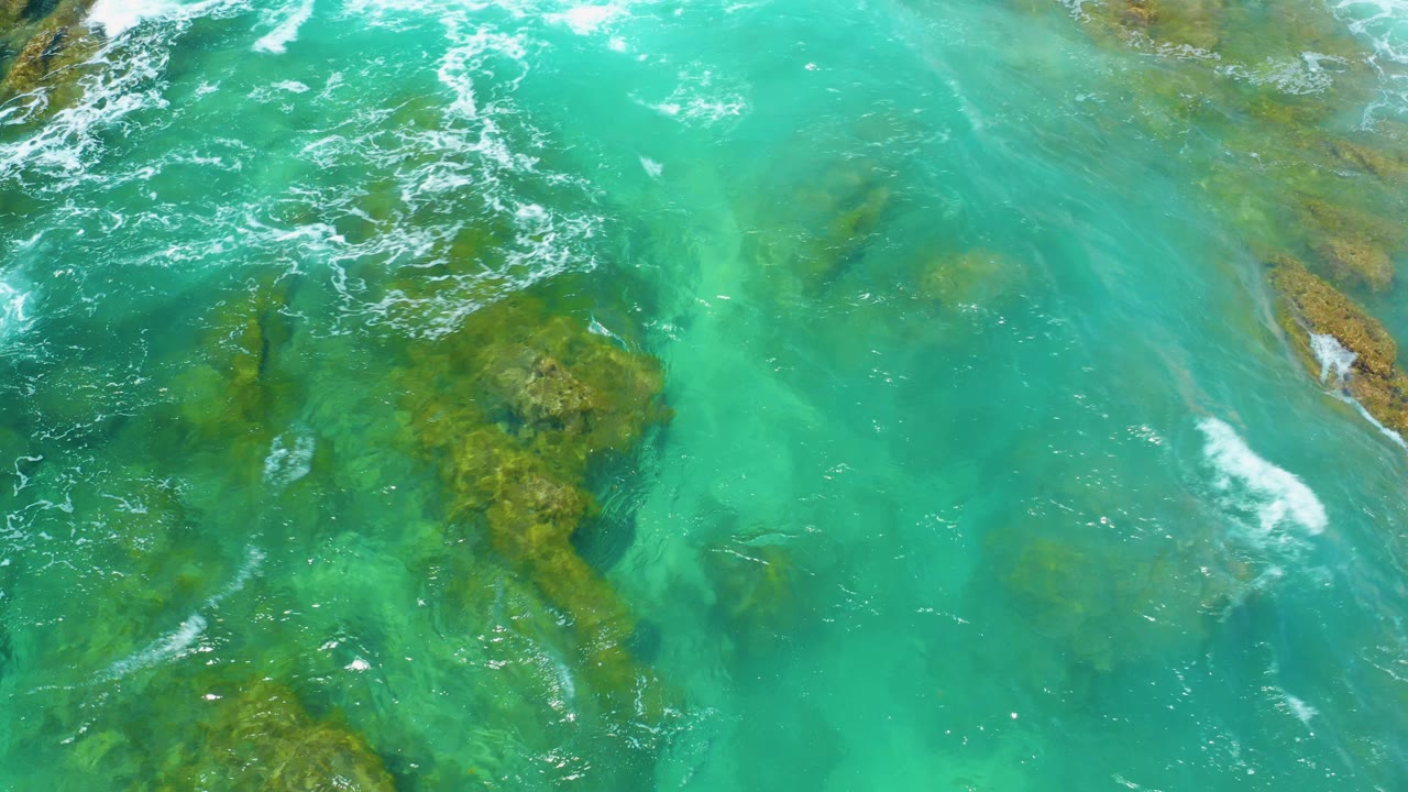 Overhead view of turquoise water at Shipwrecks Beach, Baja California Sur, with submerged rock patches, white foam lines near the upper edge, and no visible people.