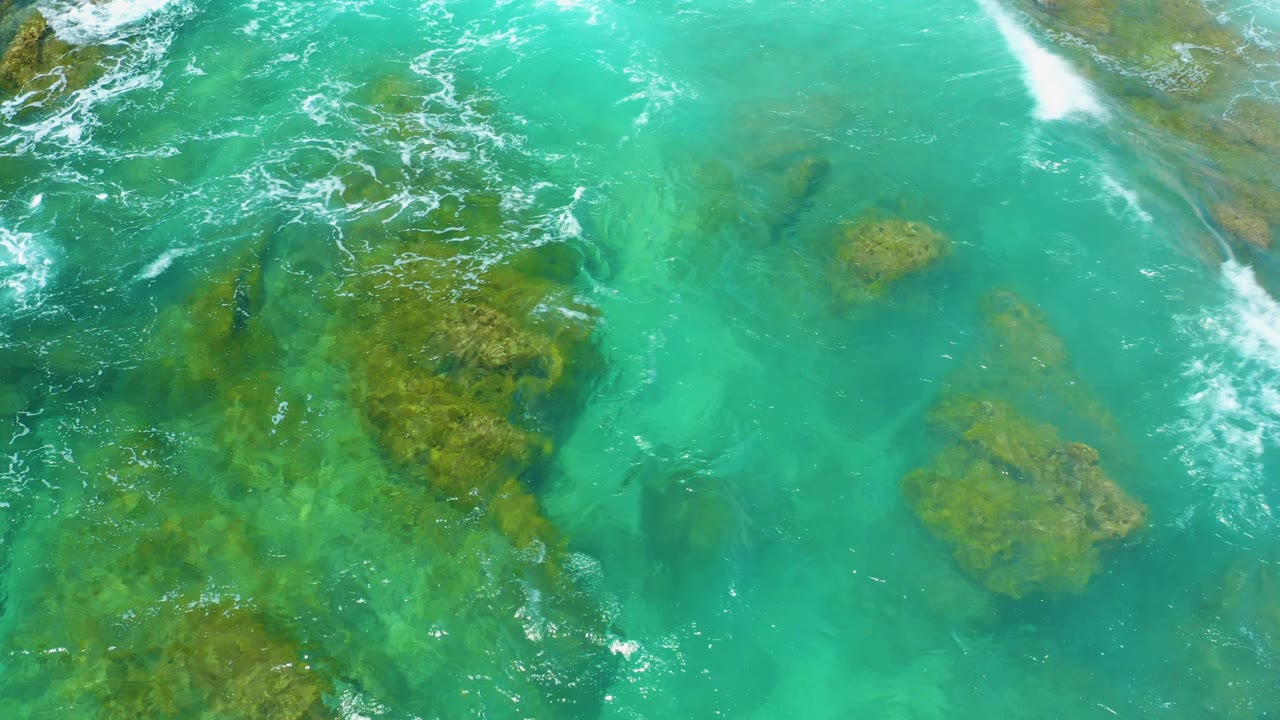 Overhead Shipwrecks Beach water view with larger brown-green rock formations under clear turquoise water, broken foam at the top, and no visible people.