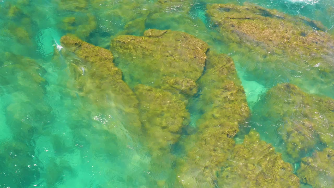 Clear green-blue water covers uneven coastal rocks at Shipwrecks Beach, with small bright foam marks and no visible people.