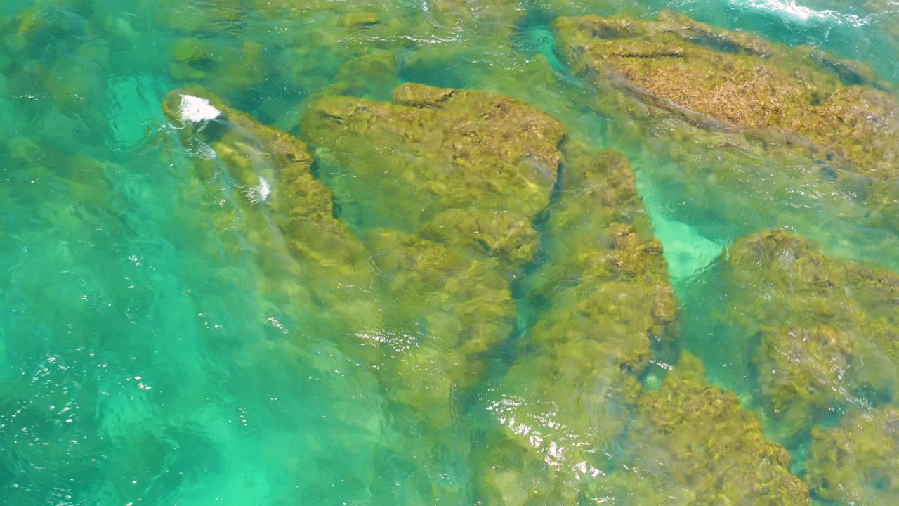 White surf enters the overhead frame near the upper rock edge at Shipwrecks Beach while turquoise water fills the surrounding shallows, with no visible people.