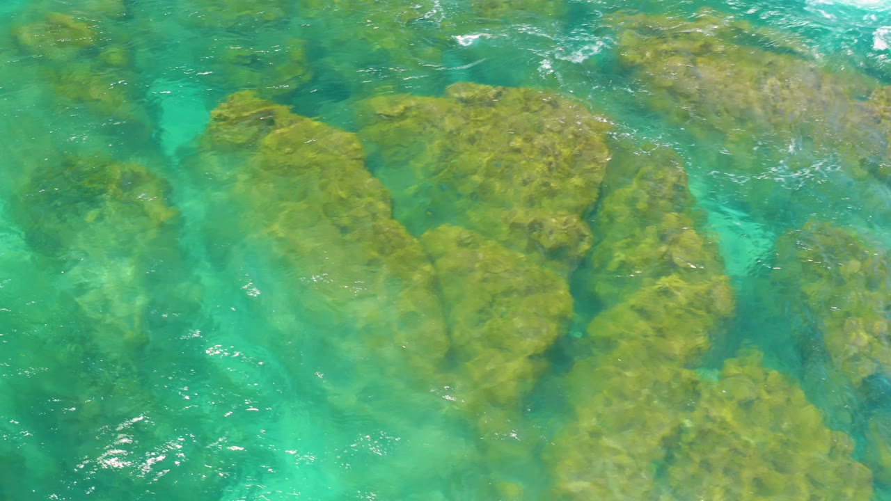 An overhead frame shows submerged rock ledges under bright green-blue water, with soft surface texture and no visible people.