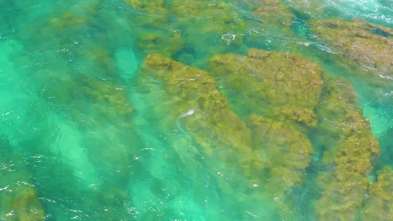 Aerial frame of green-blue water sliding over clustered shoreline rocks at Shipwrecks Beach, with pale sand tones visible below and no visible people.
