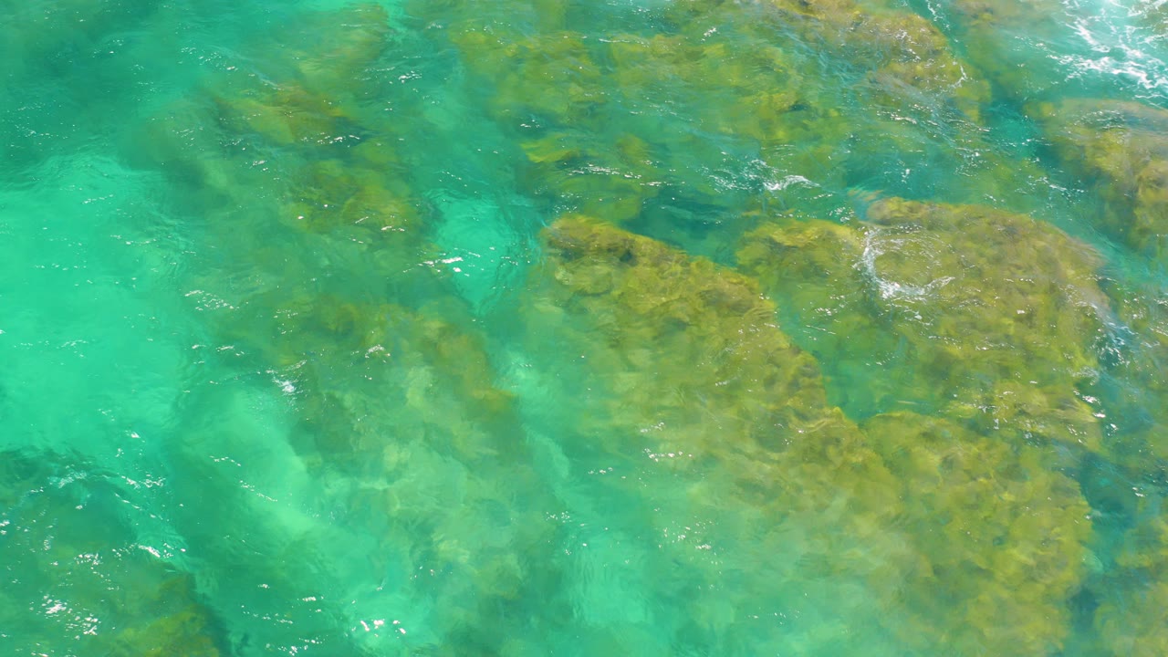 At Shipwrecks Beach in Baja California Sur, clear turquoise water covers tan submerged rocks and pale sandy patches, with no visible people.