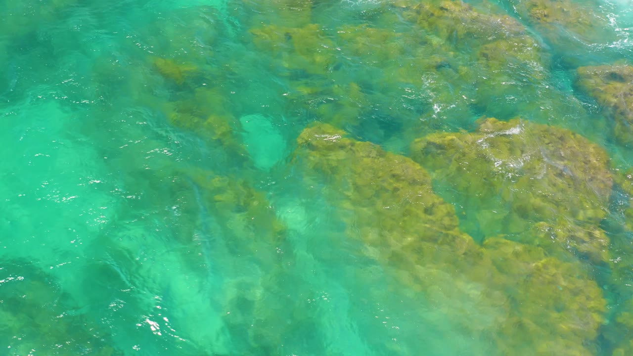 Shallow turquoise water at Shipwrecks Beach reveals broad rock shapes under the surface, with faint white foam near the upper edge and no visible people.