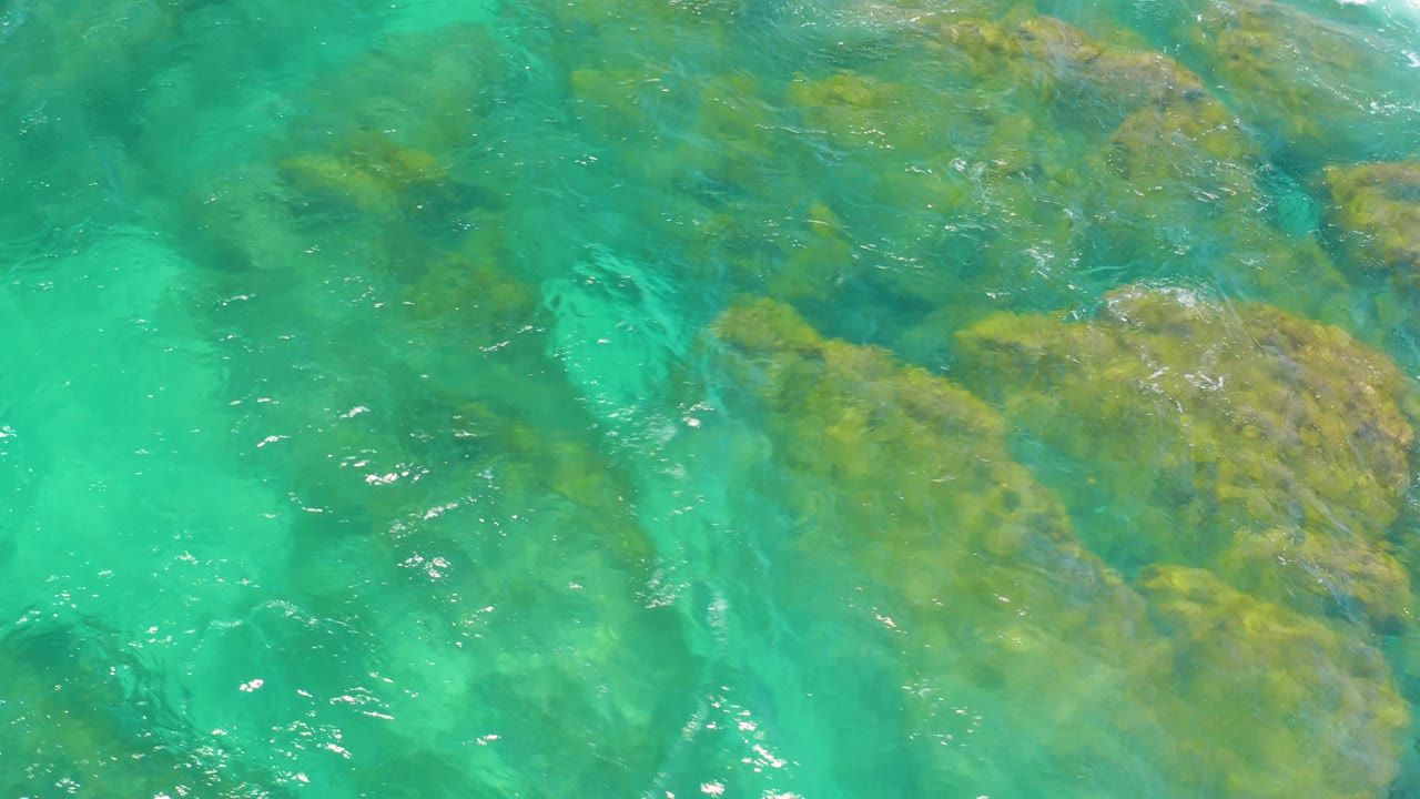 Bright green-blue water washes over clustered rocks at Shipwrecks Beach, with a soft veil of surf crossing the left side and no visible people.
