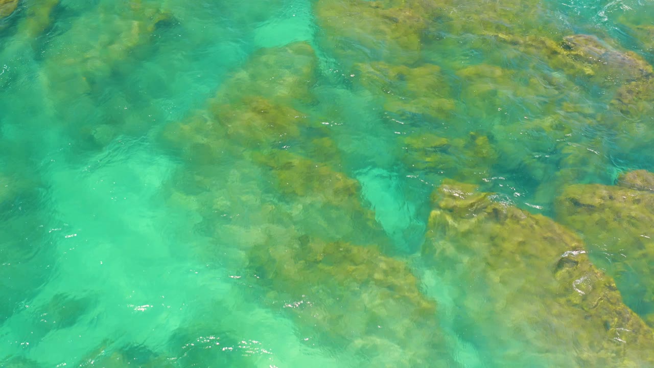 Clear green water swirls around darker submerged rocks and lighter sandy-looking patches at Shipwrecks Beach, Baja California Sur, with no visible people.