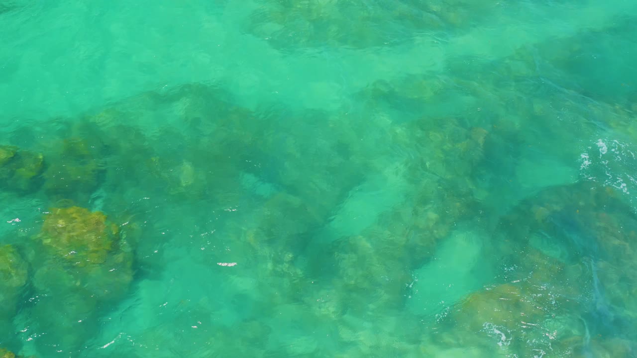 Green-blue water at Shipwrecks Beach shows pale sandy patches between darker submerged rock shapes, with fine surface shimmer and no visible people.