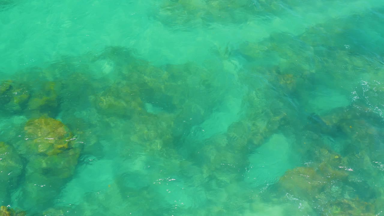 Overhead view at Shipwrecks Beach with bright turquoise water, algae-covered rock clusters, and soft wave movement across the surface, no visible people.