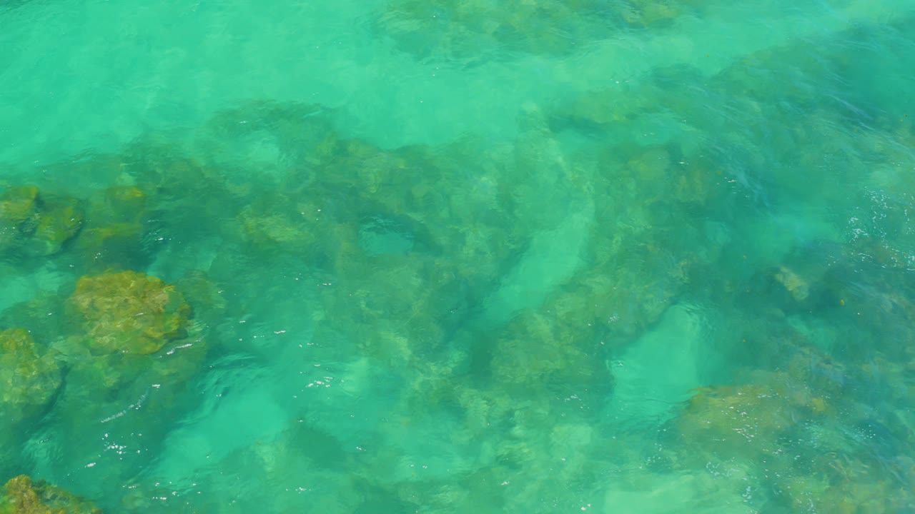Shallow turquoise ocean at Shipwrecks Beach fills the frame as soft wave bands pass over darker rocks and sandy bottom, with no visible people.